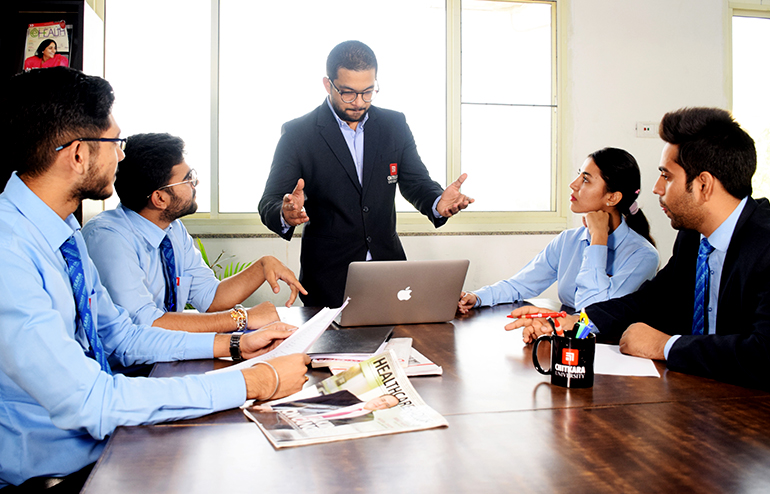 The illustration shows the students of chitkara university. One is standing, and the others are sitting around the table. A newspaper, laptop and black mug are on the table with a chitkara university logo.