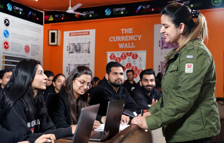 The illustration shows the classroom of chitkara university, where students sit at a desk with laptops in black sweatshirts, and the teacher teaches them.