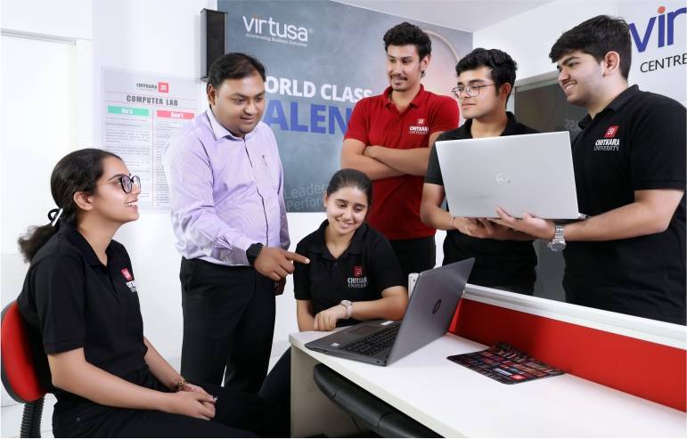 The image with a blurred background with a chitkara university logo shows the students and teacher of chitkara university—some in black t-shirts sitting on the chairs, some standing and holding the laptop. Others in red t-shirts are standing, and the teacher is standing and pointing towards the laptop on the table.