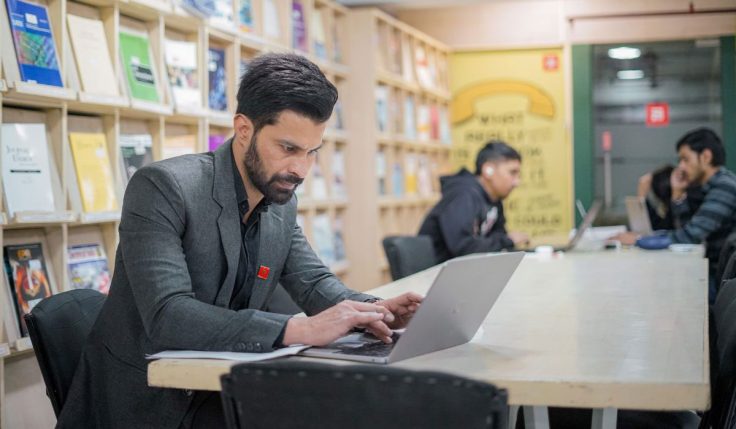 A focused student working on a laptop in a university library, engaged in research for PhD in Architecture & Urban Design studies.